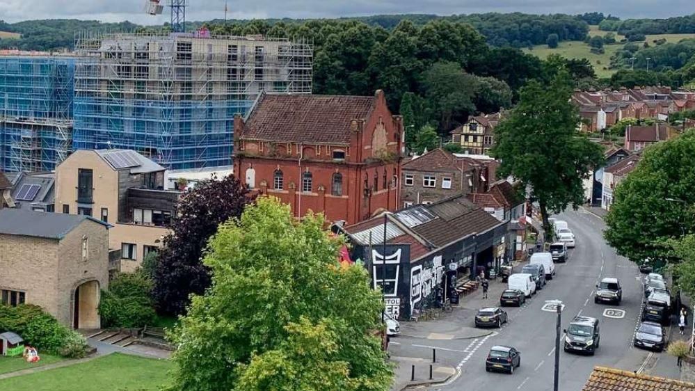 Bristol Beer Factory. The brewery is a black building with white branding on it. Other buildings are around it. A road can be seen next to the brewery, which has both parked and moving vehicles on it. 