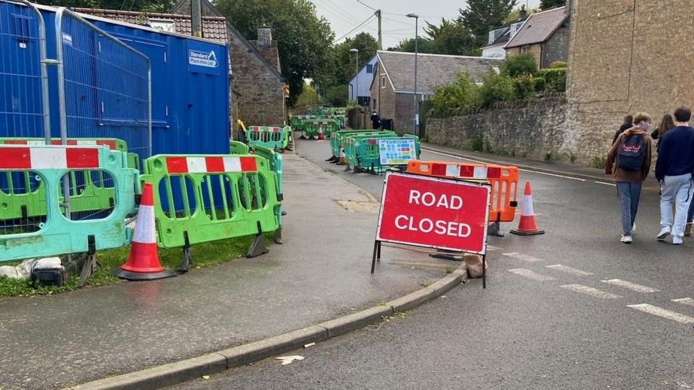 A group of teenagers walking past a 'road closed' sign and green barriers along a road