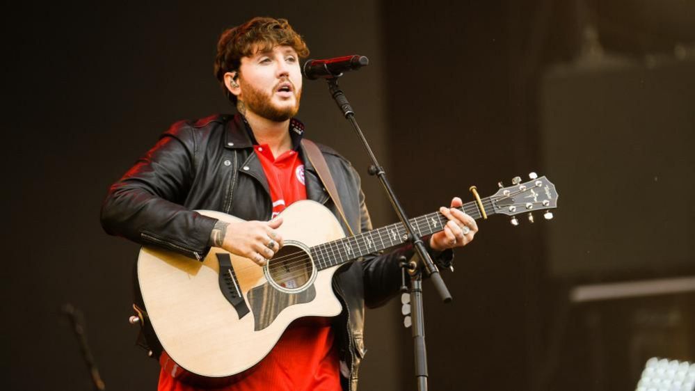 James Arthur with short brown hair and beard playing a guitar on stage