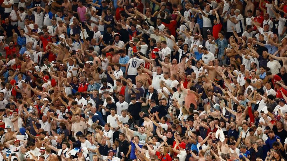 A crowd of England fans visible in the stands after the game with the Netherlands. Many in the largely male crowd have their hands raised in celebration, with others having no shirts on.