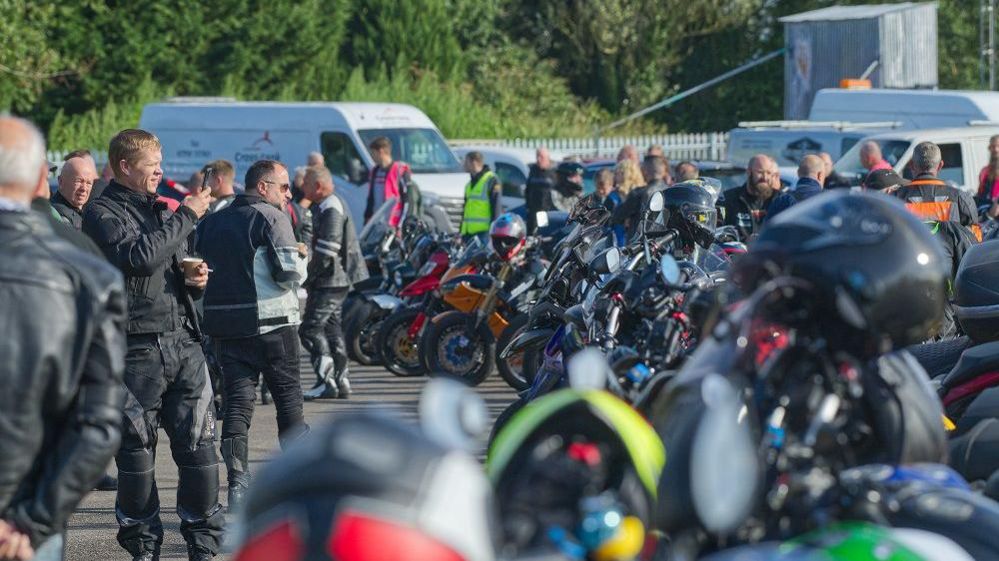 A row of motorbikes with motorcyclists in full gear milling around them