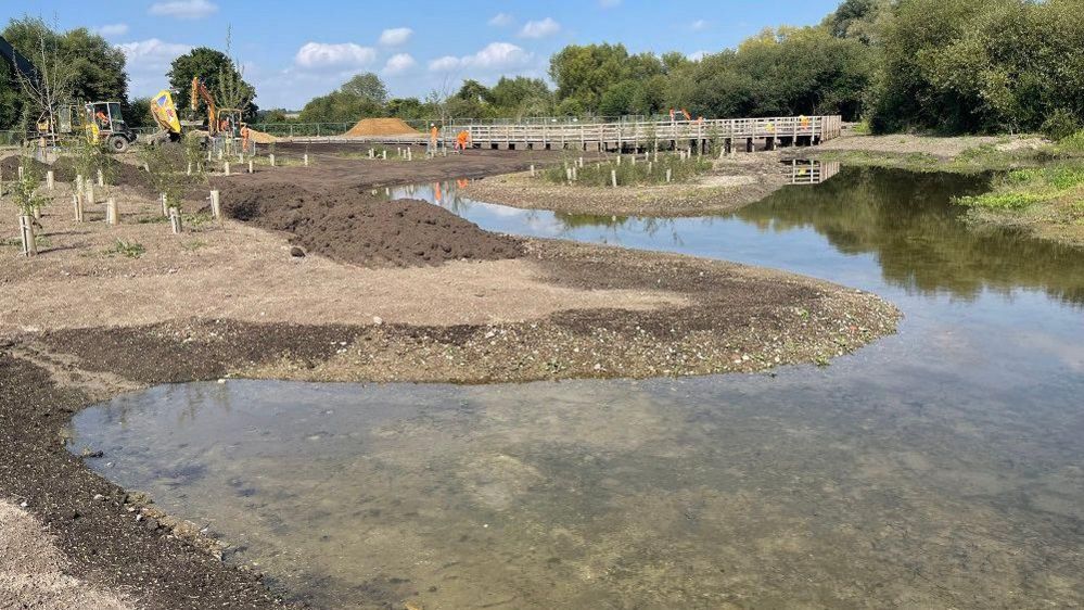 A meandering river with large banks and a couple of diggers in the background