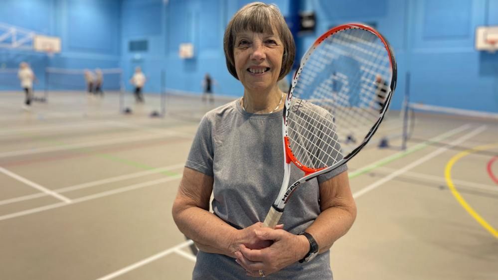 Sue Gill stands with a tennis racquet in a sports hall