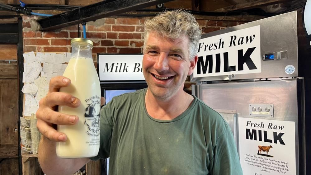 Robin Shreeve holding a bottle of raw milk next to the vending machine in his farm shop