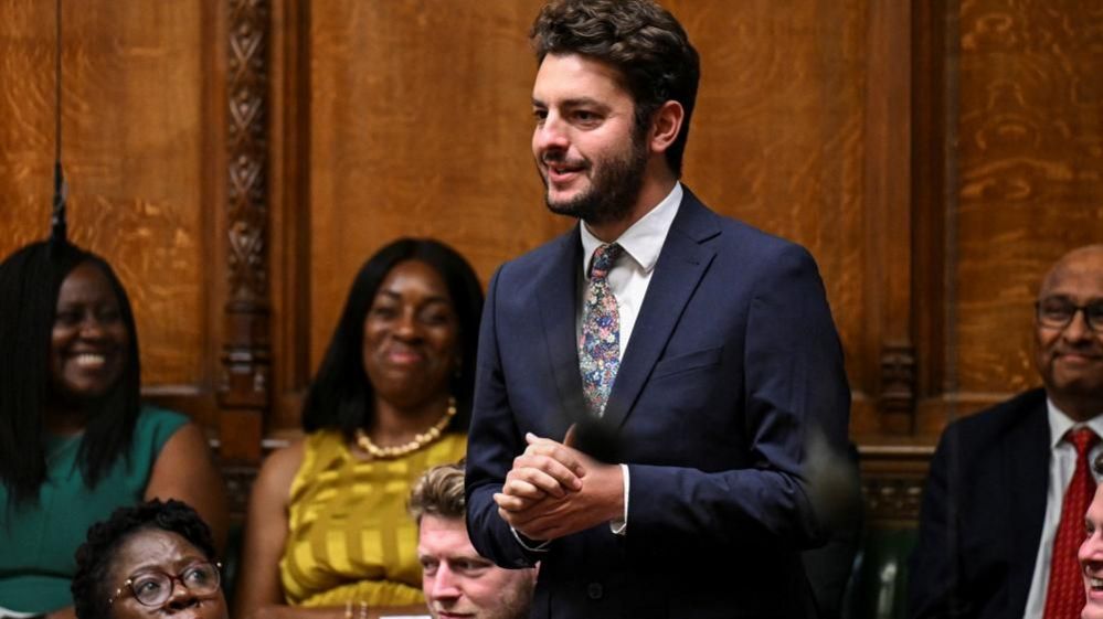 MP Jack Abbott standing in House of Commons with other MPs looking on. He is wearing a dark suit, white shirt and and multicoloured ties with stars on it