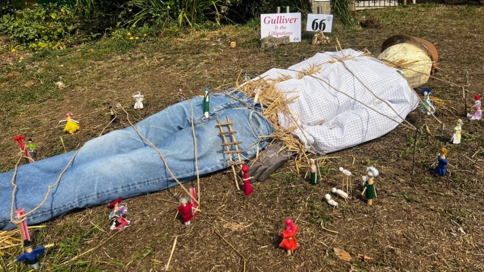 A Gulliver scarecrow, dressed in jeans and a checked shirt, wearing a hat, lying on the floor with small toys pinning him down