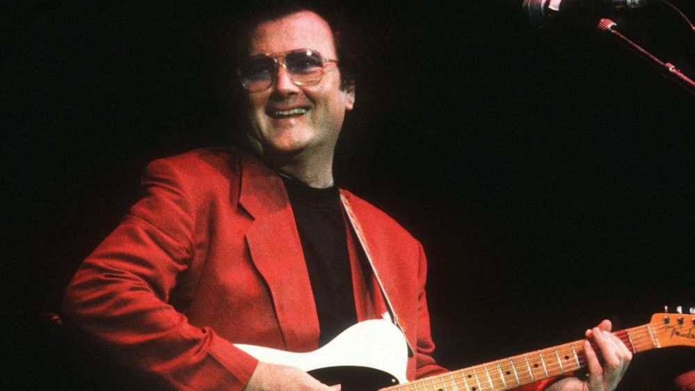 Gerry Rafferty in sunglasses and a red suit while playing the guitar