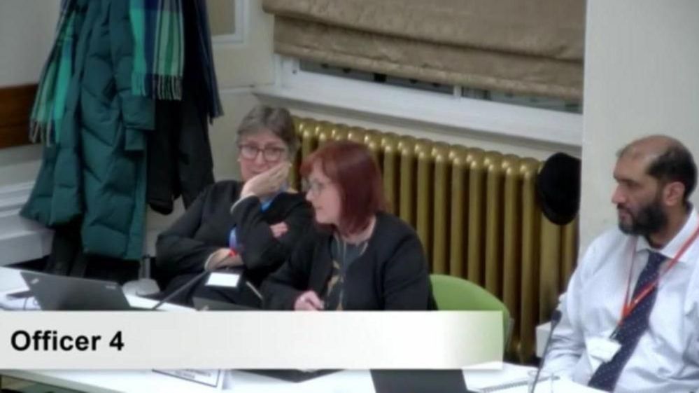 A screenshot from a council meeting picturing two women and one man seated at desks with microphones. A woman with red hair and glasses is speaking.