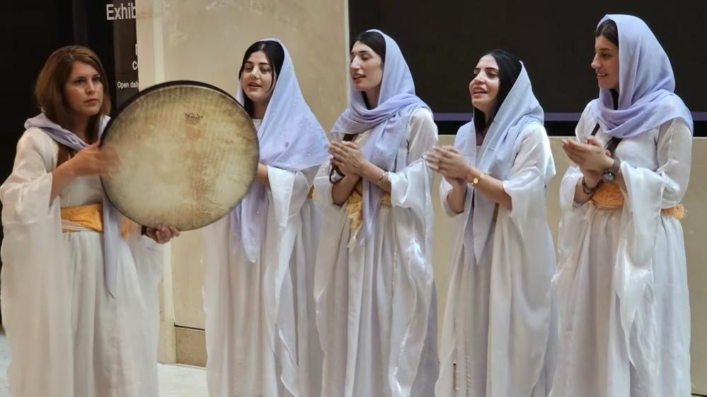 A group of women in robes and headscarfs, one holding a drum, sing