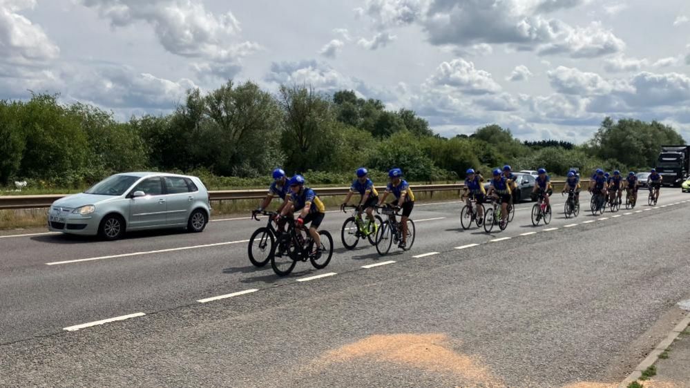 A line of bikes in the middle lane of a three-lane road. All riders have blue tops and helmets