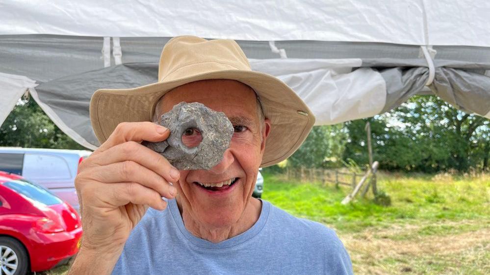 Peter Warry holds an artefact in front of his eye and to the camera to demonstrate a "spyhole"