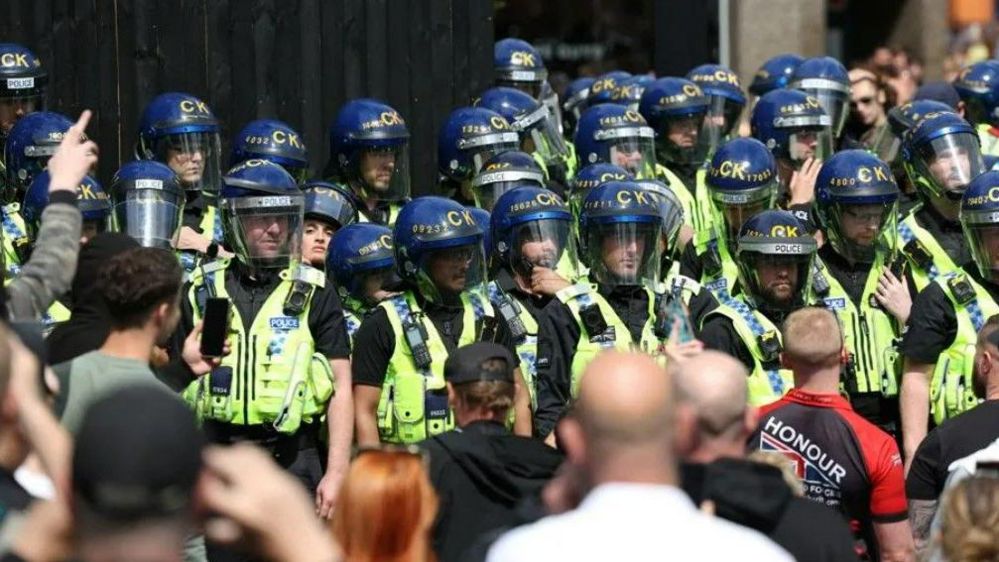 Police officers in riot gear stand guard as people participate in a protest in Manchester