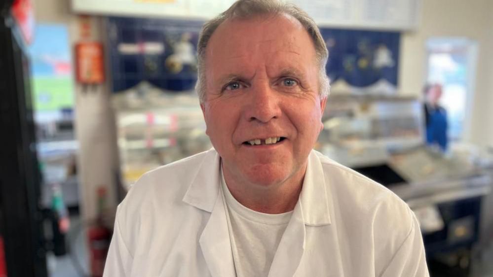 Peter McGowan wearing a white overall and T-shirt with a fish fryer in his fish and chip shop blurred in the background 
