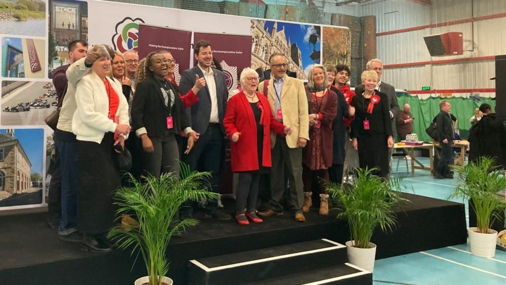 Danielle Stone wearing a red top and standing with supporters on a stage