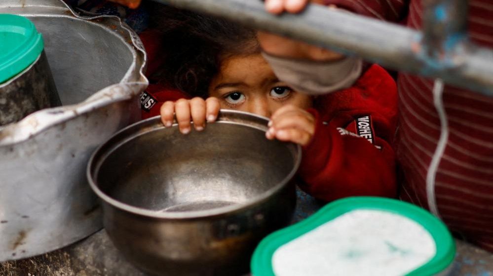 A young child peeks over the rim of an empty bowl she holds while standing in a crowd awaiting food aid in Rafah