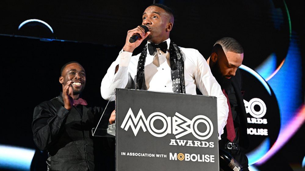 Bugzy Malone at the Mobo Awards in Sheffield. Bugzy wears a black bow tie and neck scarf over a pressed white shirt.