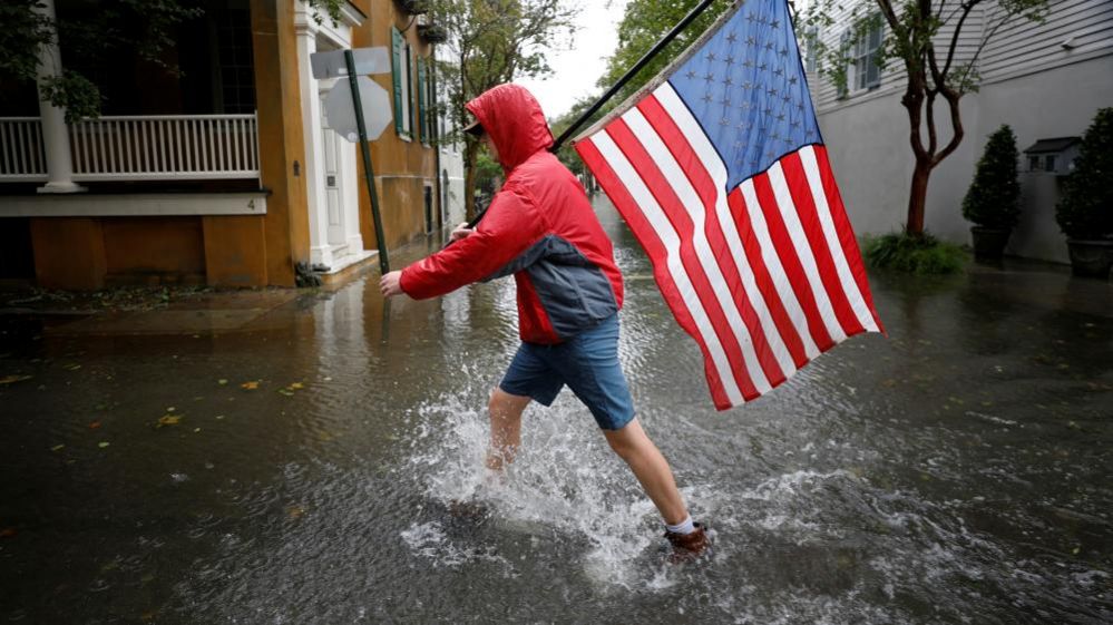 A man walking carrying a flag in a flooded Charleston street