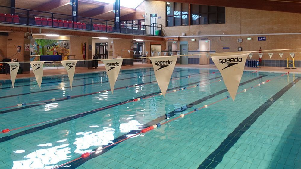 An indoor swimming pool with lanes. Across the pool is white branded bunting. There is a balcony with a of seats 