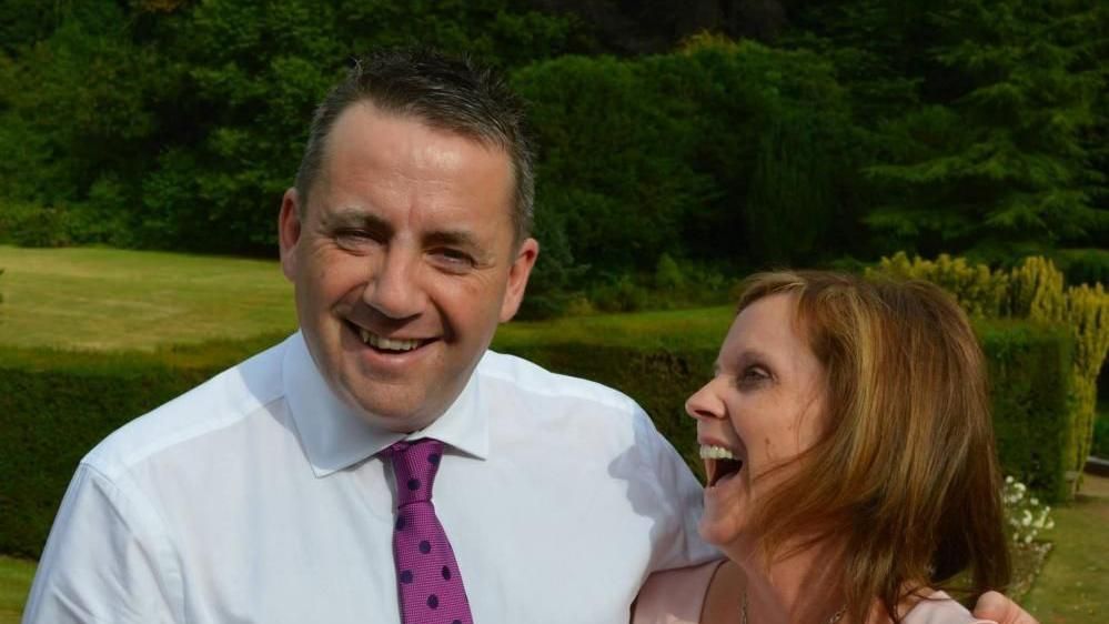 John Hunt dressed in a white shirt and pink-spotted tie smiling at the camera with his arm around his wife Carol, who is laughing and looking at her husband