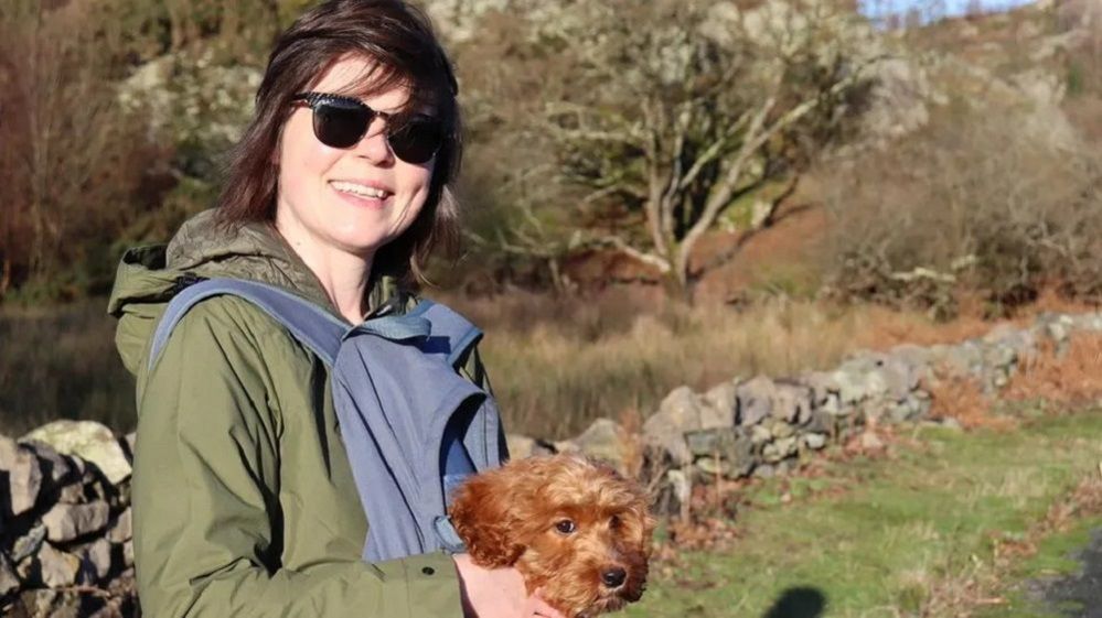 Lowri, in sunglasses, carrying a brown dog