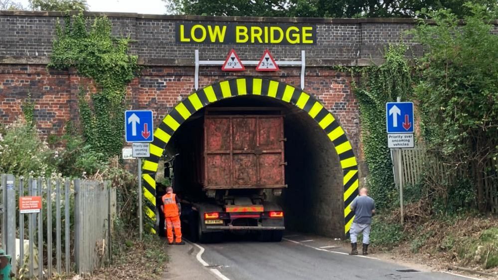 Back of a red lorry is visible inside a bridge under the sign "Low Bridge". A man in orange overalls and another man in a grey shirt are looking at the lorry.