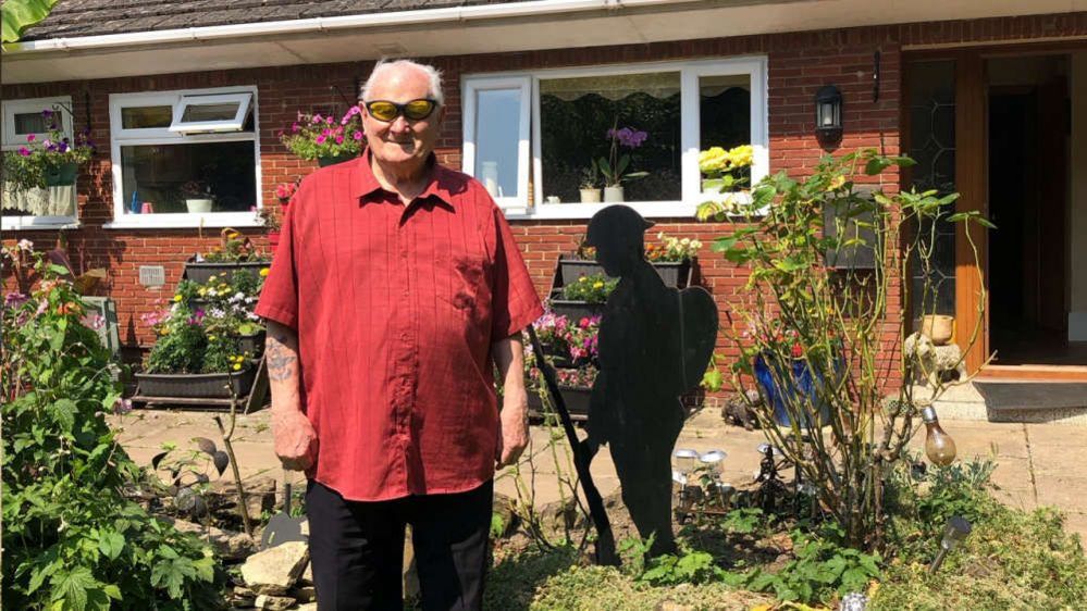 Danny Gibbon wearing a red shirt, yellow tinted glasses and black trousers. He is standing in his garden beside lots of flower beds and a silhouetted statue of a soldier. 