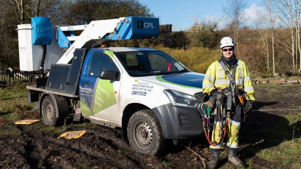 An Electricity North West engineer wearing safety equipment. He is standing in a muddy field with a cherry picker behind him.