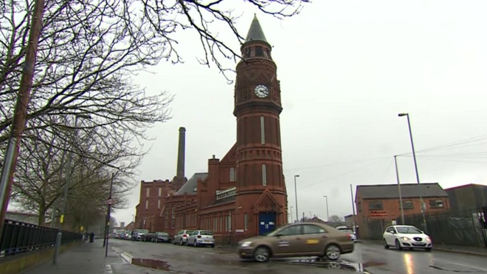 Front view of the mosque with a tall clock tower and the red-brick building behind it along the street with cars parked outside