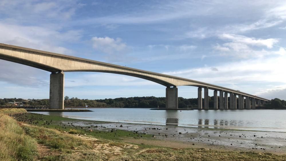 A view of the Orwell Bridge from a bank by the side of the River Orwell. The bridge is large, grey structure with a river running beneath it. 