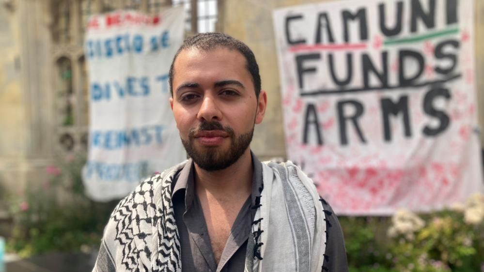 Mahmoud Atallah, who has short hair and a beard, stands in front of placards at the camp