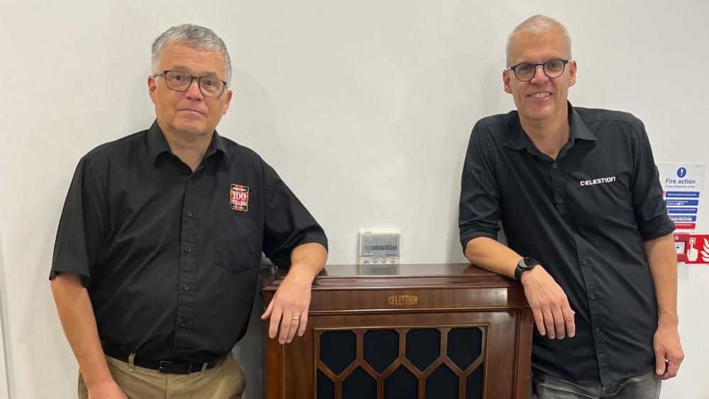 Two men wearing glasses and black shirts are leaning on a wooden piece of furniture.