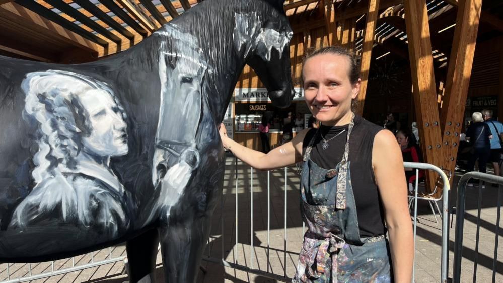 Lois Cordelia standing next to the partially-completed painting on a model of a horse