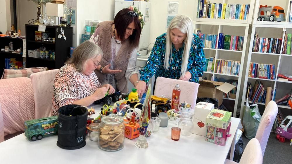 Three women around a table on which are toys of various kinds. Susannah has long blonde hair and is wearing a black and blue dress