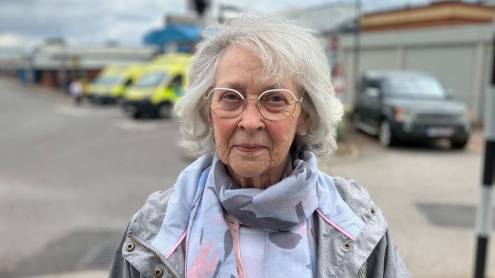 Jill Henderson with white hair wearing a grey raincoat with ambulances in the background