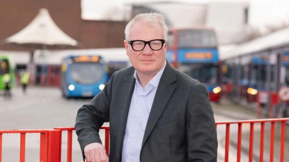 Richard Parker leaning on a red metal railing in front of a blurred background of a bus station