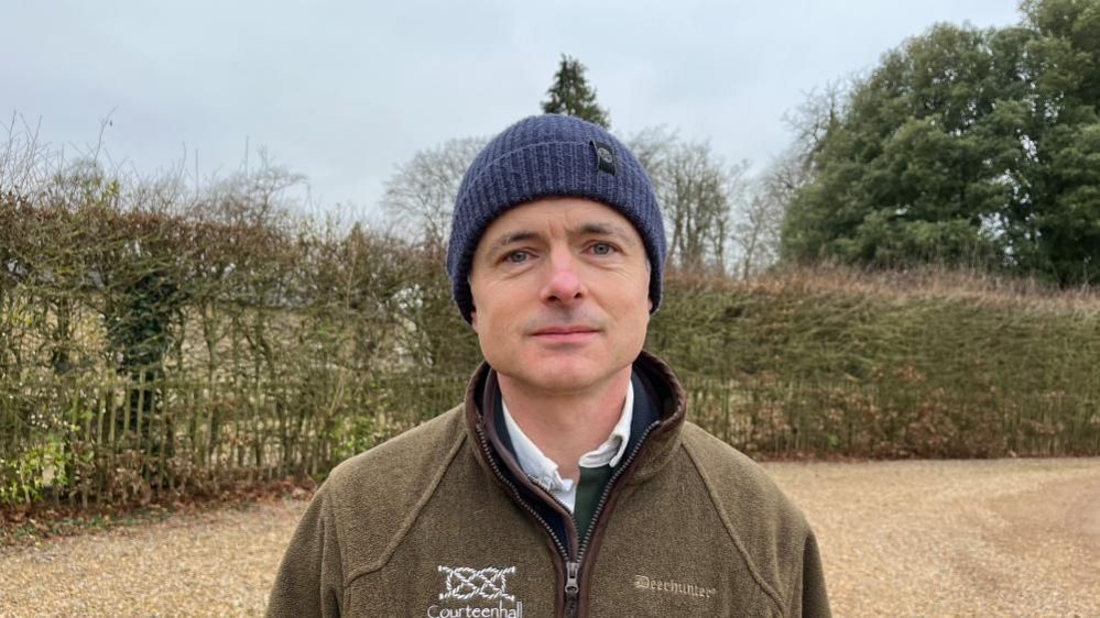 Johnny Wake wearing a green jacket and blue bobble hat standing on a gravel drive next to a hedge