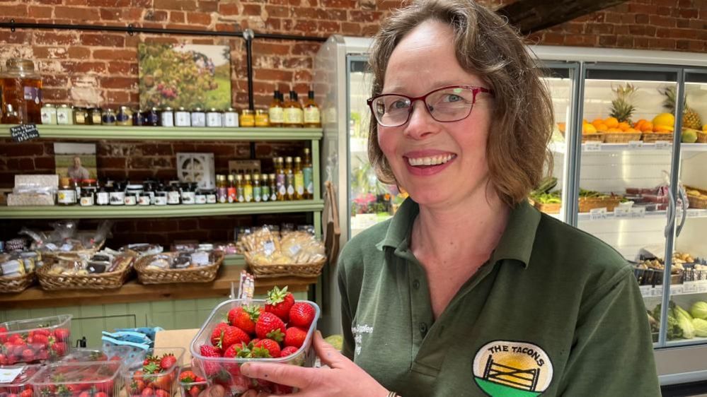 A smiling Emma Tacon holding up a punnet of strawberries, while standing in her shop