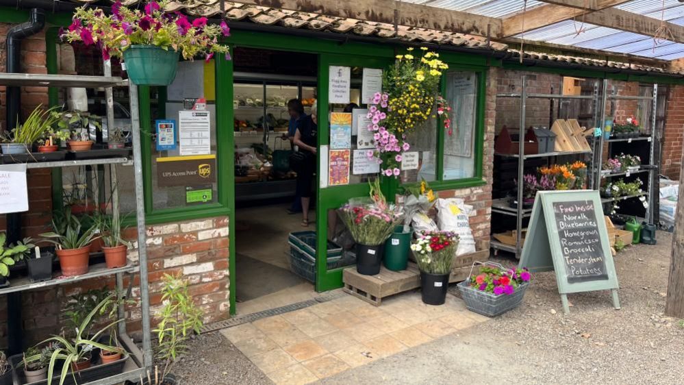 Entrance to The Tacons' farm shop, a single-storey red brick building with flowers and plants outside 