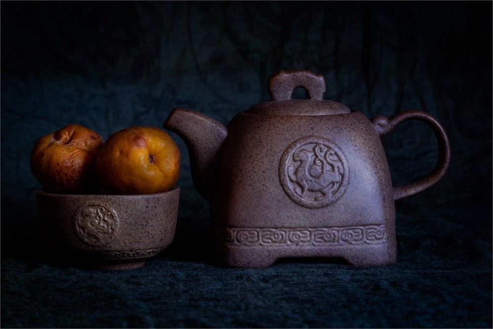 A black teapot with a dragon motif on it next to a fruit bowl with matching motif that has quince in it