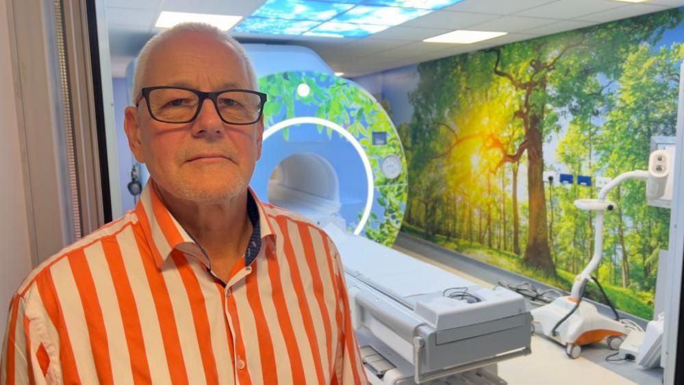 Colin Horn, wearing a bright orange and cream striped top, standing by a scanner that has been decorated with nature scenes