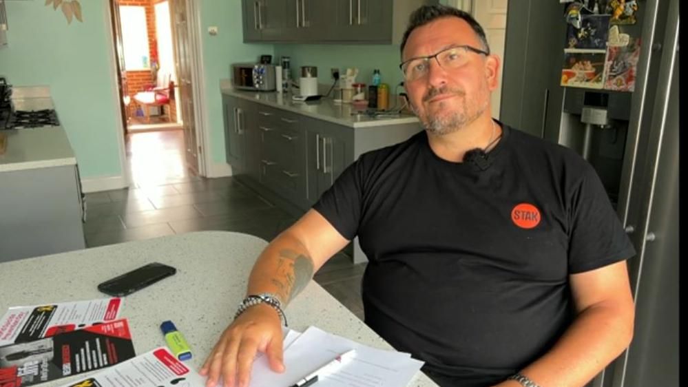 Tristan Kluibenschadl sits at his kitchen table wearing a black t-shirt with the Stak logo on it