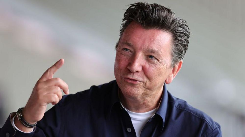 Nigel Le Quesne in a blue shirt smiling to the left of camera with his index finger pointed upwards