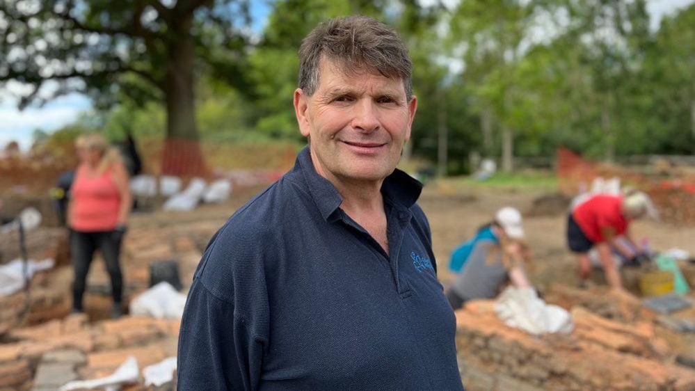 Neil Holbrook wearing a blue polo shirt looks into the camera from the dig site.