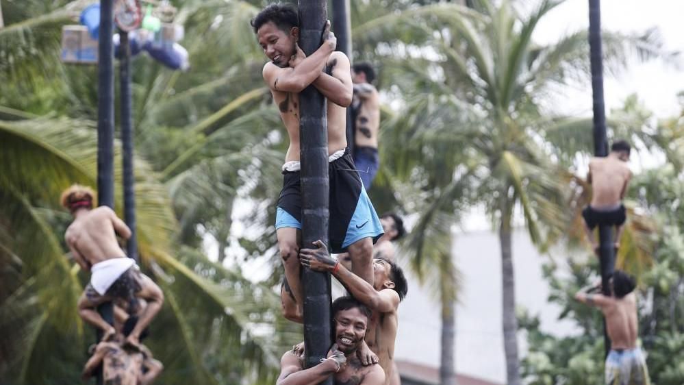 Men climb greasy poles in a race. Some stand on each other's shoulders against a backdrop of palm trees. 