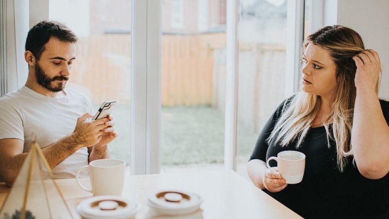 Casal sentado diante da mesa: ele olha para o celular, e ela parece incomodada