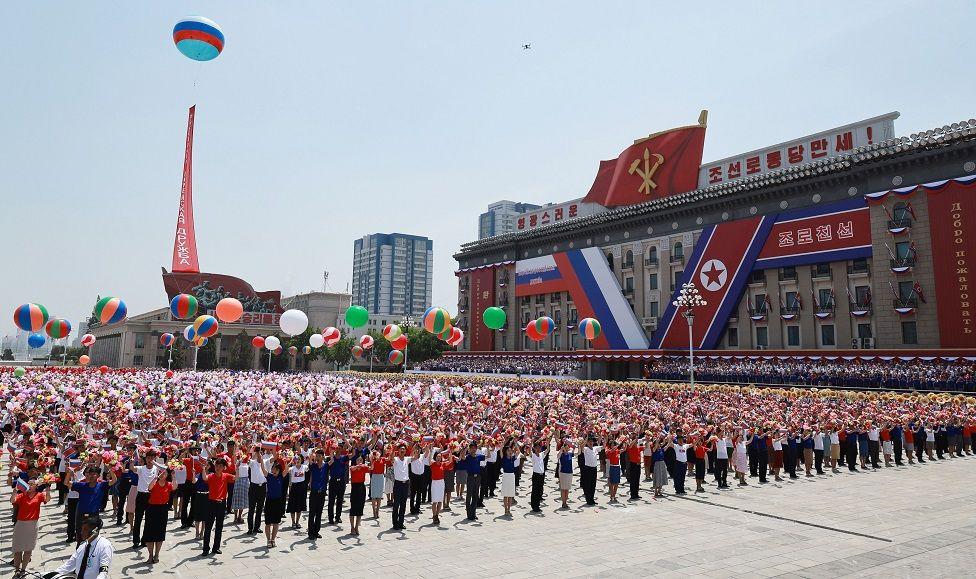 Una multitud parada con globos de colores, de un lado de la plaza se ven las banderas de Corea del Norte y Rusia
