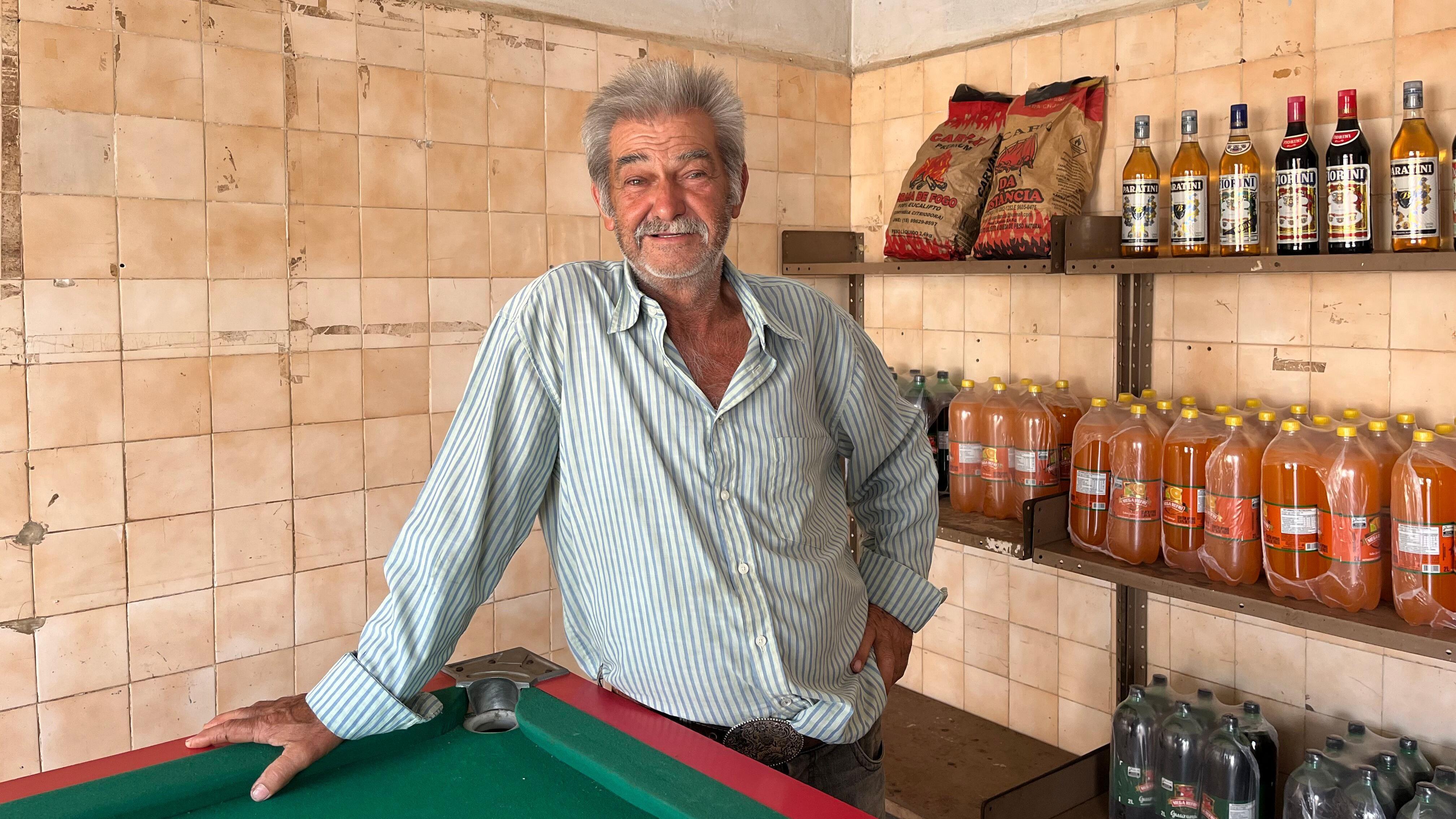 Homem sorrindo, apoiado em mesa de sinuca, dentro de bar; atrás dele, há prateleiras com bebidas