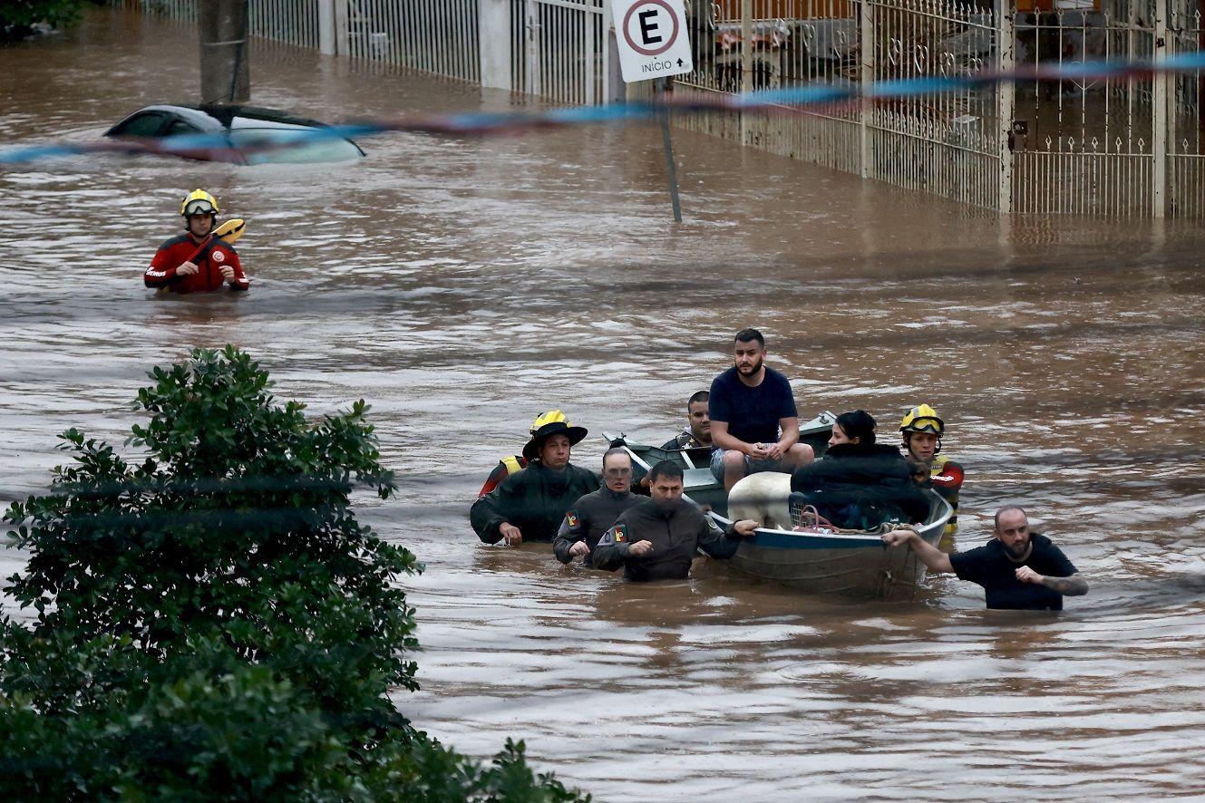 Pessoas dentro de barco, acompanhados de socorristas perto da embarcação em rua inundada