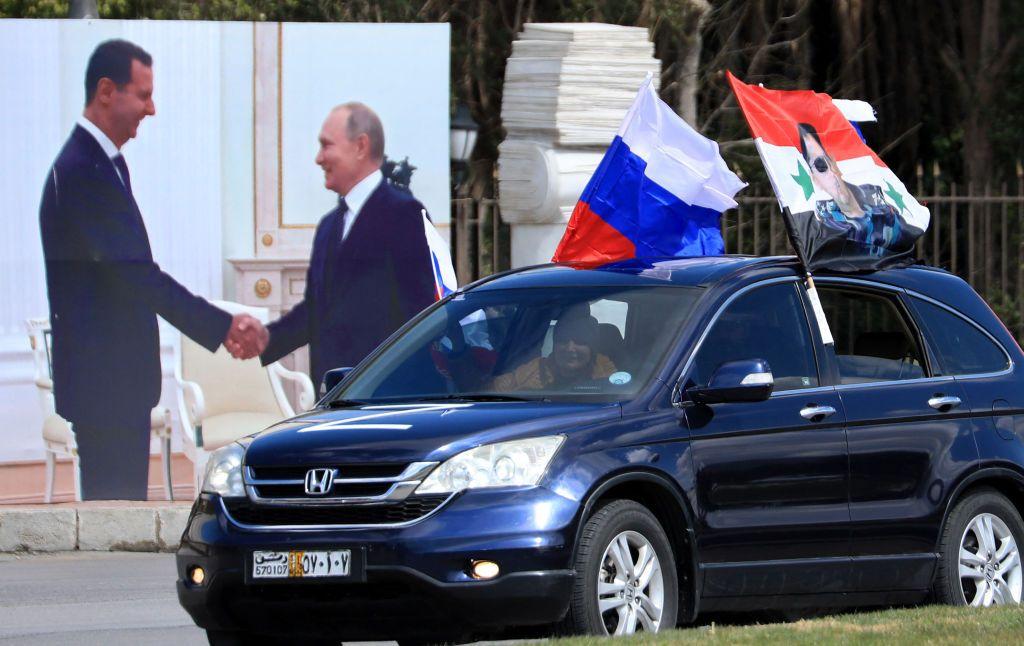 Un auto con bandera rusas y sirias pasa por una pantalla que muestra a los mandatarios de ambos países en 2022.