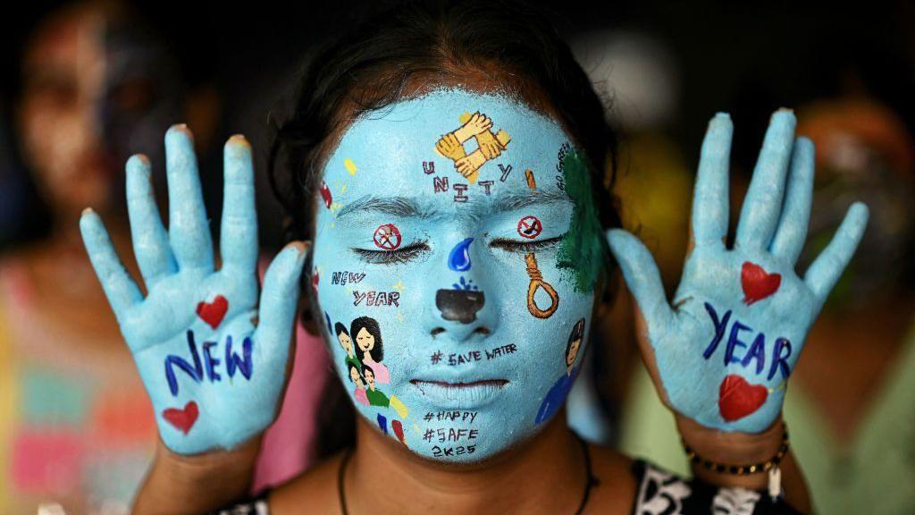 Foto de um rosto pintado de azul com desenhos e uma mão de cada lado, com as palmas para a câmera, também pintadas de azul, com os dizeres 